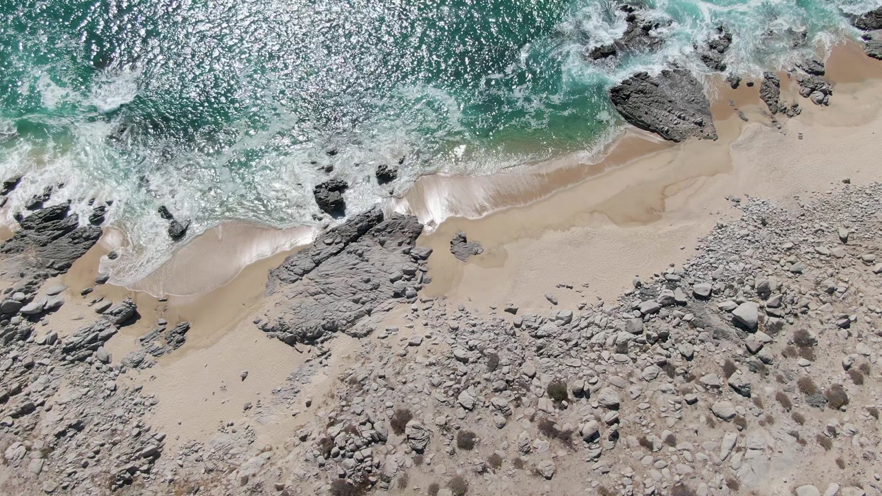 Aerial frame from Buzzards Beach showing brighter foam advancing across the upper shoreline while boulders and rough rock fill the lower beach, with no visible people.
