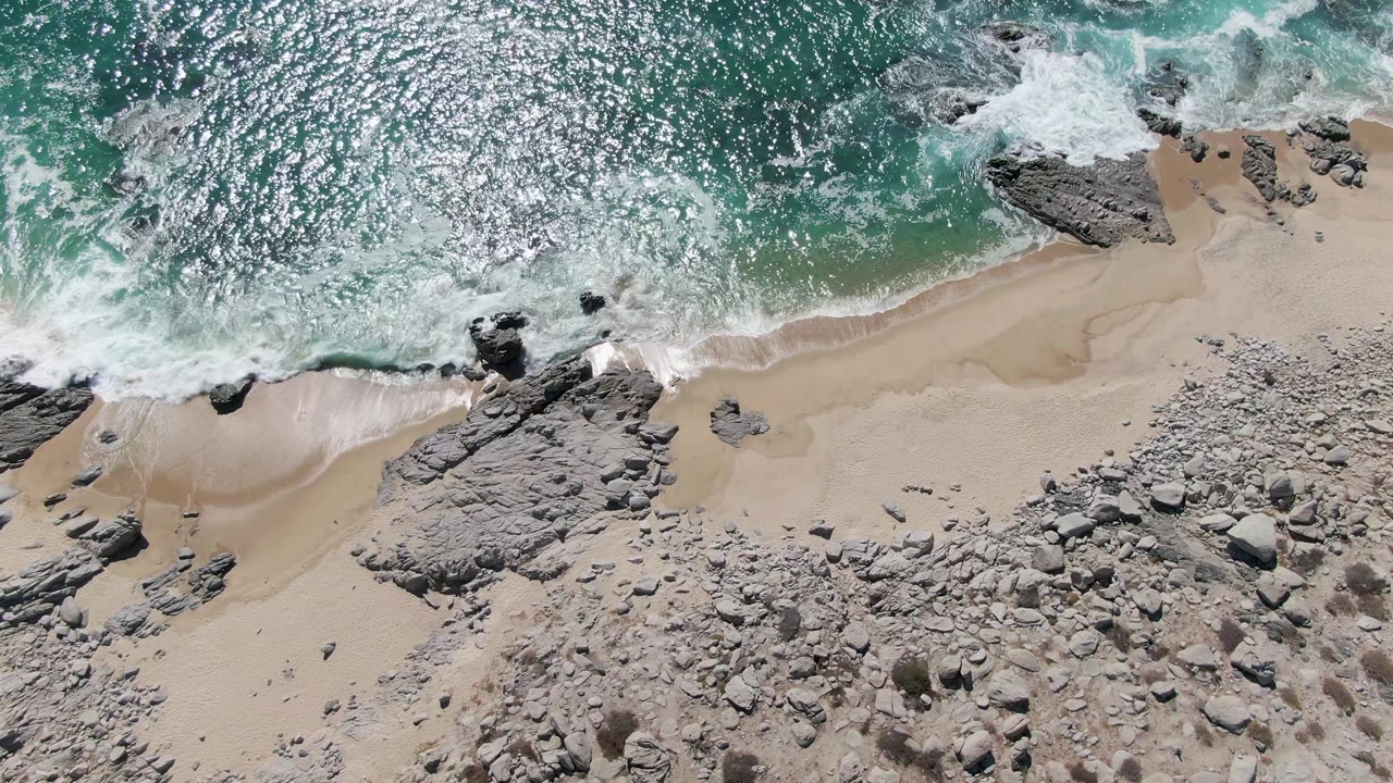 A wave spreads white foam along the midline of the Buzzards Beach shore, with turquoise water above, pale sand below, gray rock outcrops, and no visible people.