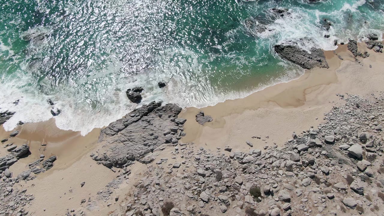 Foamy surf curls around a dark rock near the Buzzards Beach waterline, with turquoise water, pale sand, broad gray stone slabs, and no visible people.