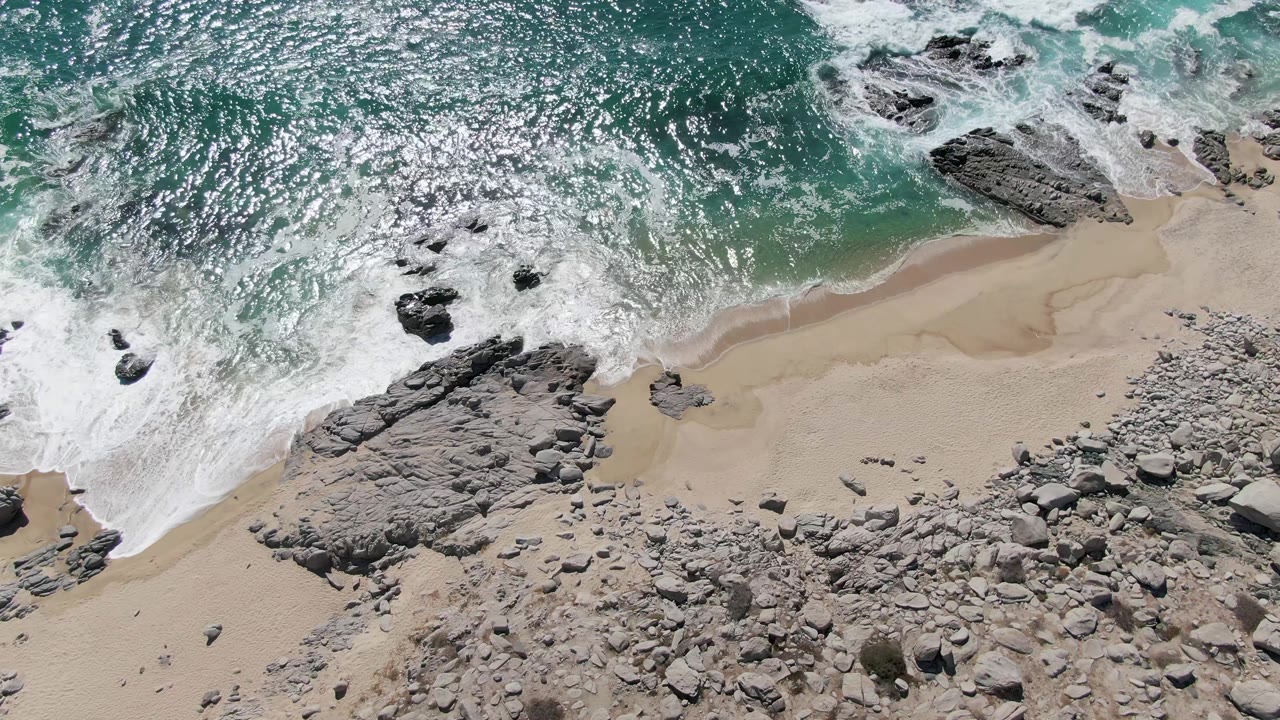 At Buzzards Beach in Baja California Sur, Mexico, foam pulls across a sandy pocket between rock formations under sparkling turquoise water, no visible people.