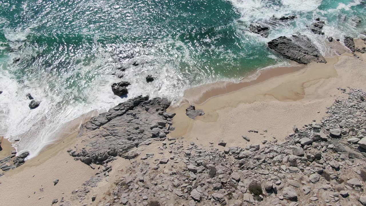Buzzards Beach shoreline seen from overhead as turquoise water breaks along the top of frame and pale sand curves around rocky outcrops, no visible people.