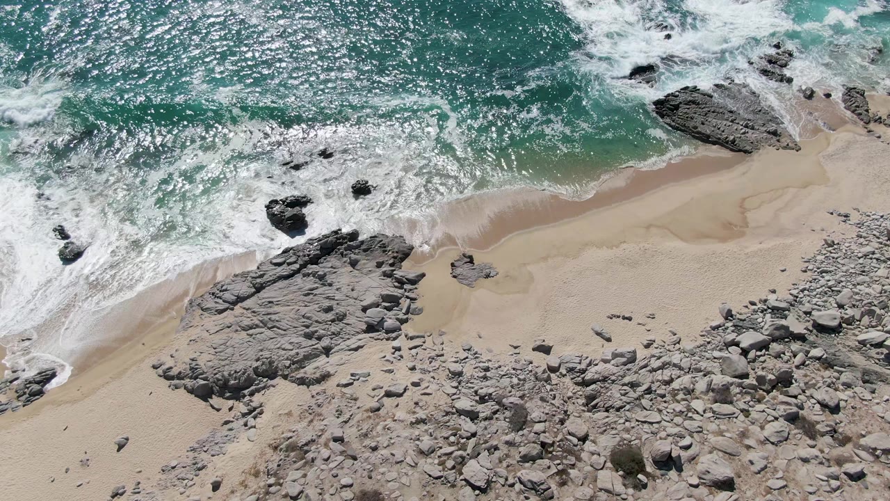 Overhead frame from Buzzards Beach in Baja California Sur, Mexico, where a foamy wave curls onto sand beside a broad gray rock shelf, no visible people.