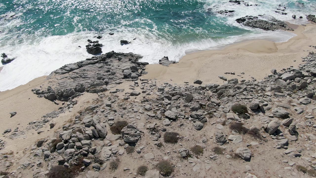 Aerial view over Buzzards Beach in Baja California Sur, with gray rocks across pale sand, turquoise surf breaking at the top, and a small dark figure near the left edge.
