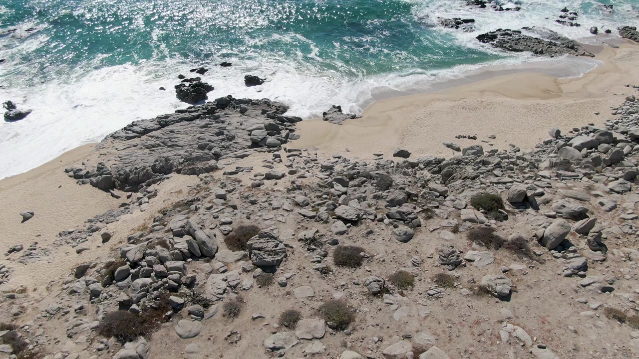 Buzzards Beach shoreline in Baja California Sur with foamy turquoise water at the top, a curved strip of wet sand, scattered rocks, and no visible people.
