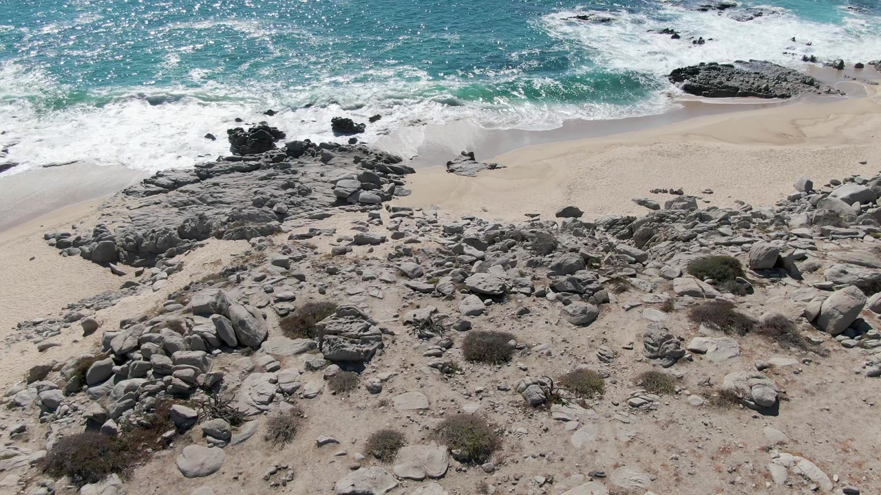 A high aerial frame at Buzzards Beach shows turquoise water breaking into white foam above pale sand and scattered gray boulders, with no visible people.