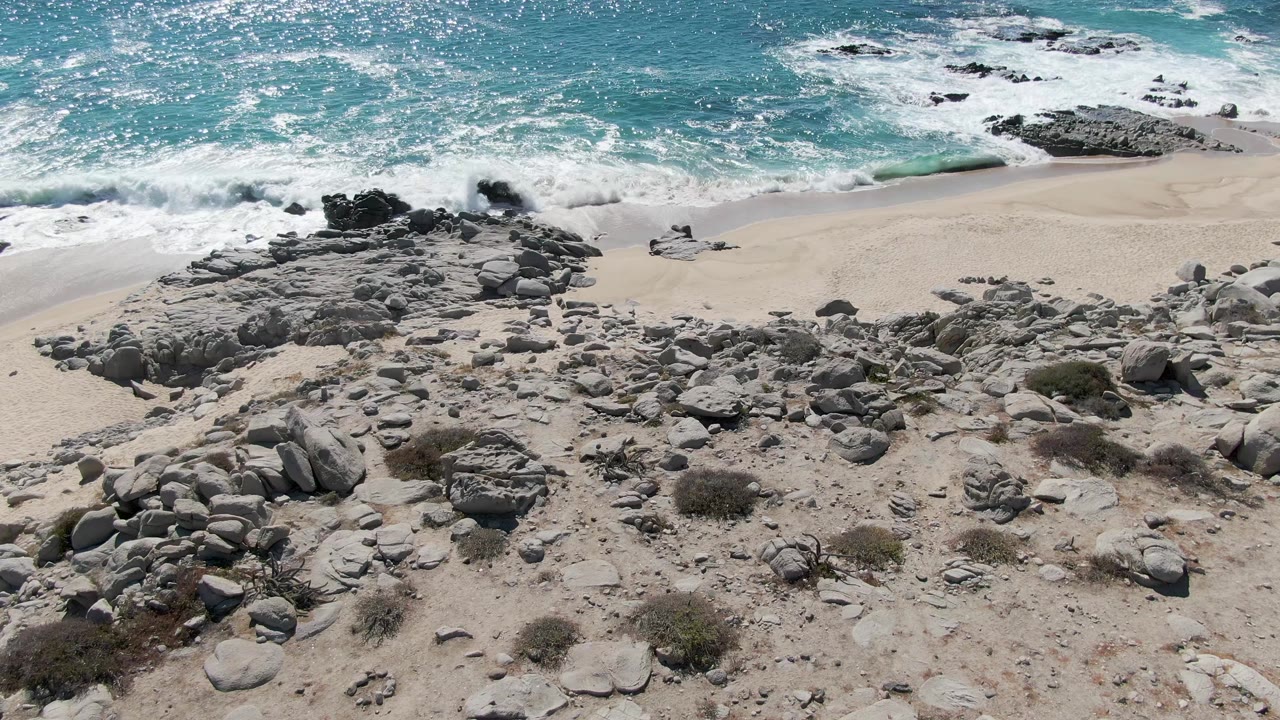 At 0:02, surf breaks around darker rocks near the waterline at Buzzards Beach, with pale sand and a broad spread of gray boulders below, and no visible people.
