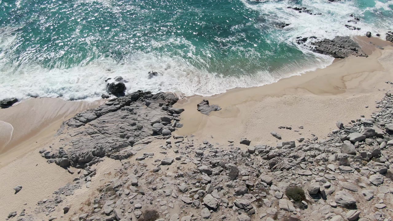 Aerial view over Buzzards Beach as white surf curls along the upper sand line near a broad gray rock shelf and scattered boulders, with no visible people.