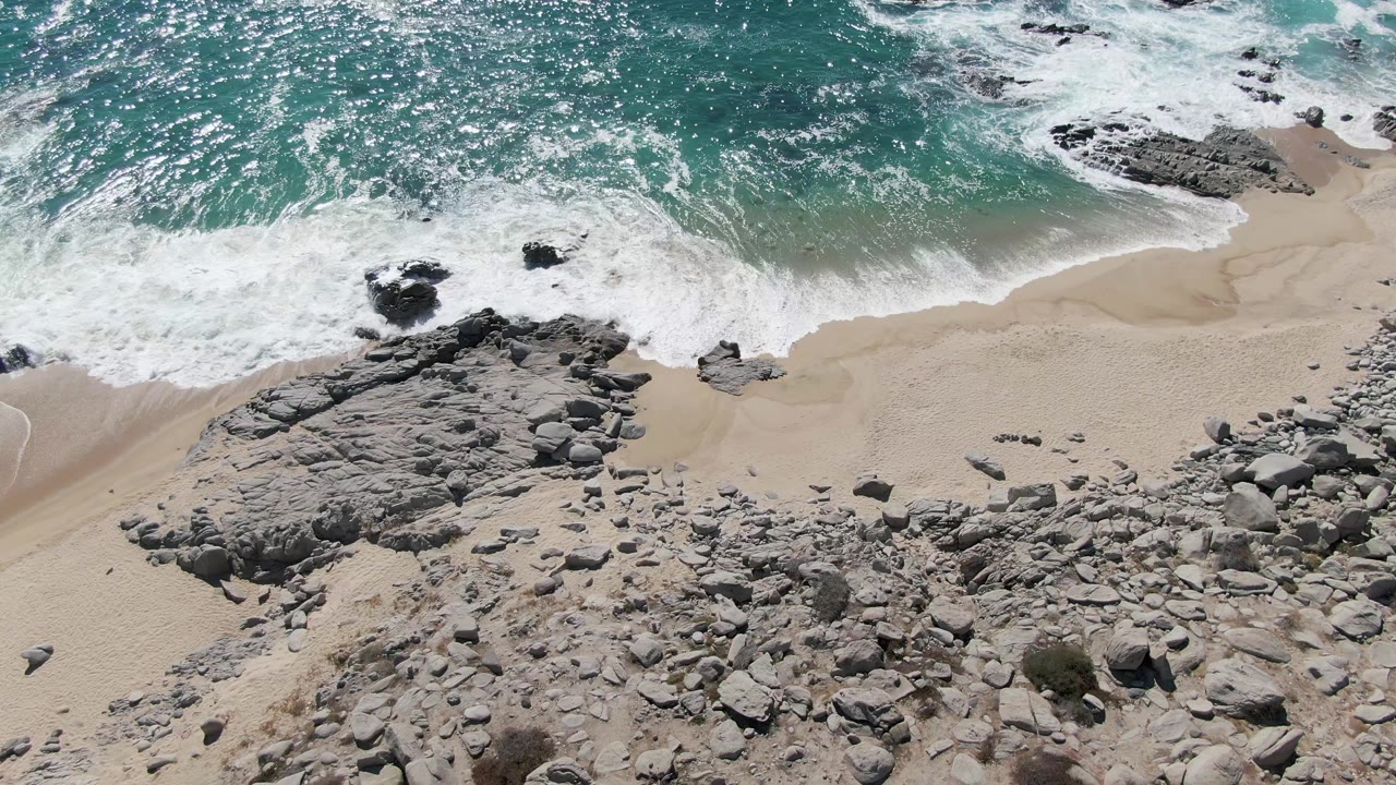 Buzzards Beach aerial frame with heavier white foam covering the shoreline, gray outcrops below it, and loose stones across the lower sand, with no visible people.