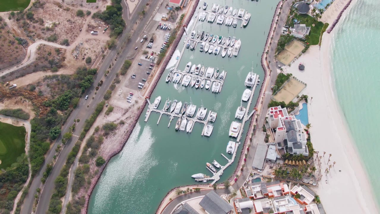 Aerial frame over La Paz, Baja California Sur showing a coastal road on the left, docked boats in the marina, sandy beach on the right, and no visible people.