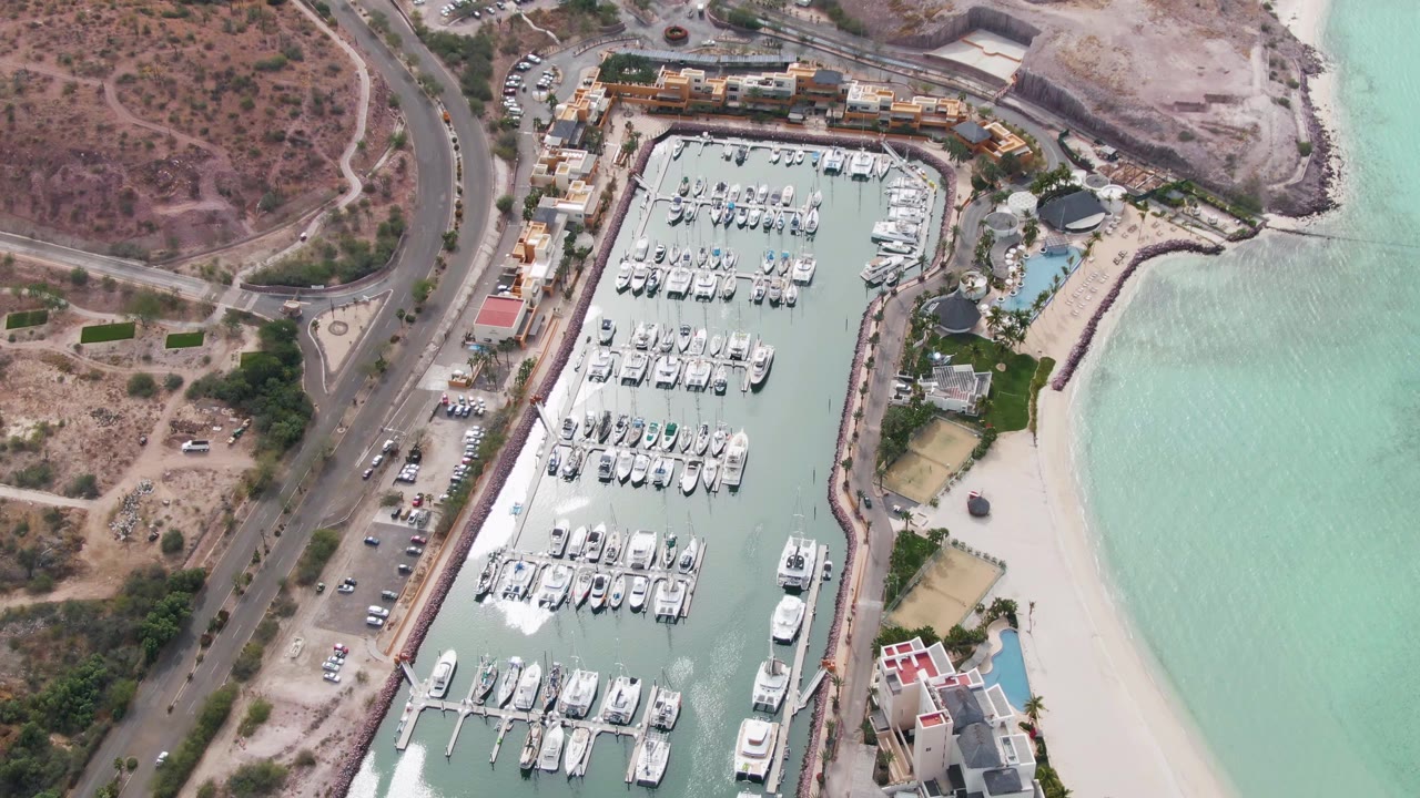 High drone frame over the La Paz marina with dock rows and boats below, dry hillside and road to the left, a curved beach and blue-green water to the right, and no visible people.