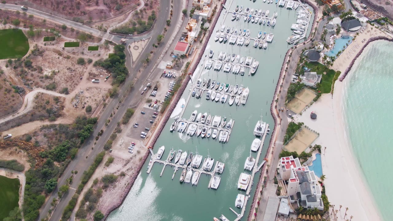 Overhead frame of La Paz, Baja California Sur showing a marina filled with docked white boats, a coastal road, pale beach, green patches, and dry hillside, with no visible people.