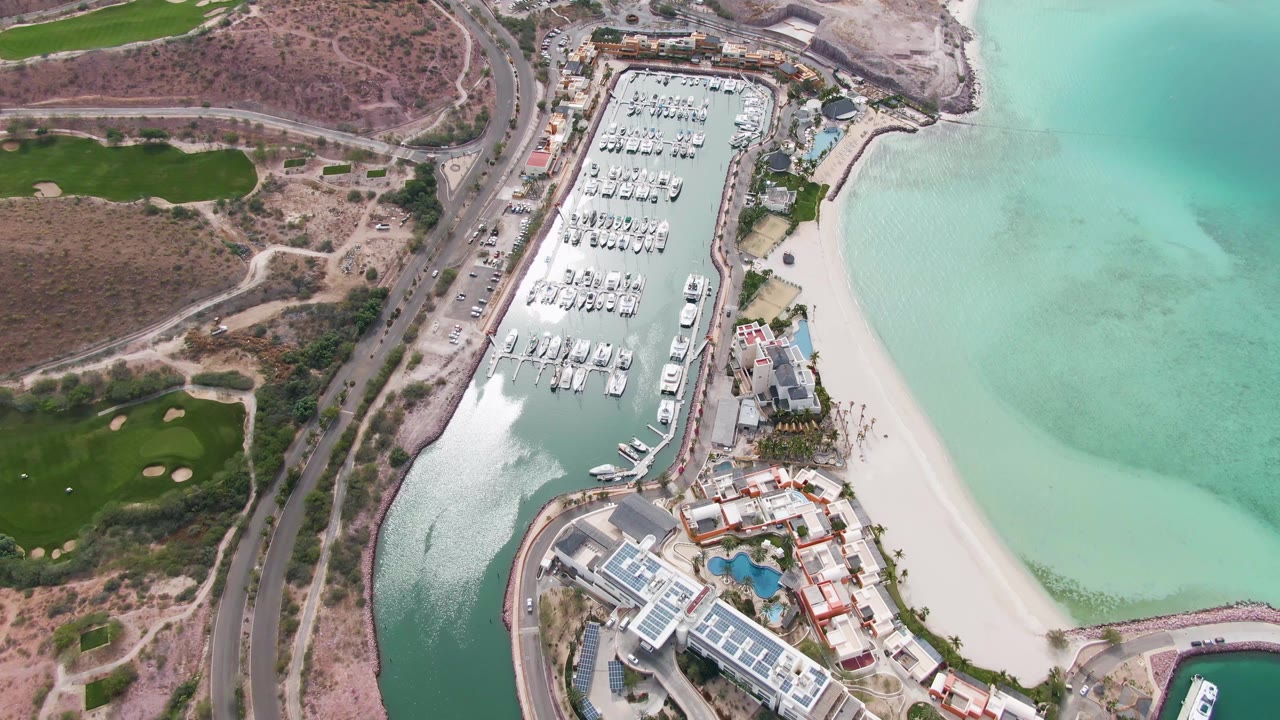 Opening aerial frame over La Paz, Mexico, with a marina packed with docked boats, a pale beach to the right, a coastal road to the left, and no visible people.