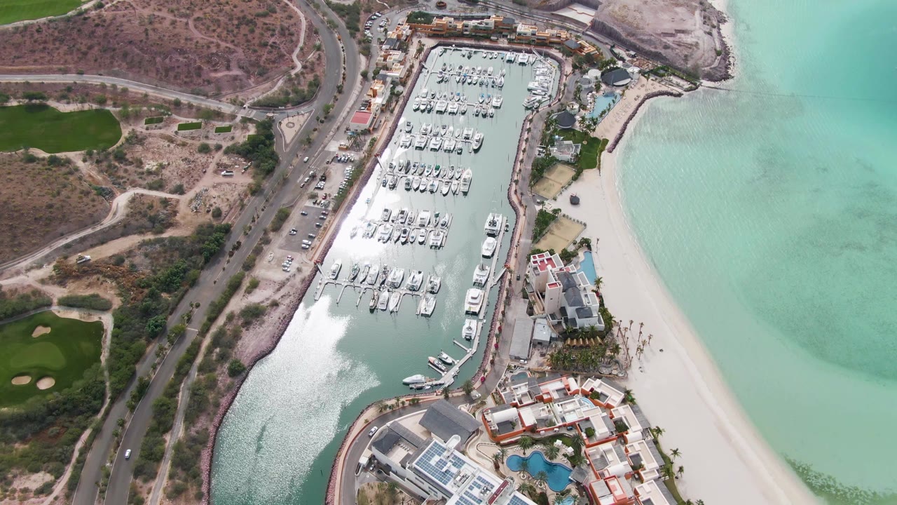 High aerial frame over La Paz, Baja California Sur, showing rows of boats in a marina, a sandy beach at right, a coastal road at left, dry hillside above, and no visible people.