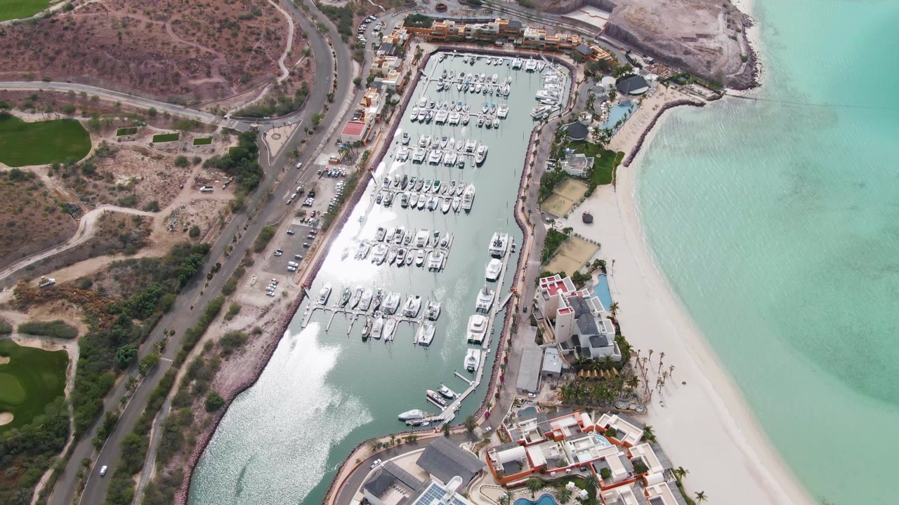 Aerial view in La Paz, Baja California Sur, with white boats arranged in slips, sunlight on the marina water, beach and shallow turquoise water to the right, road traffic to the left, and no visible people.