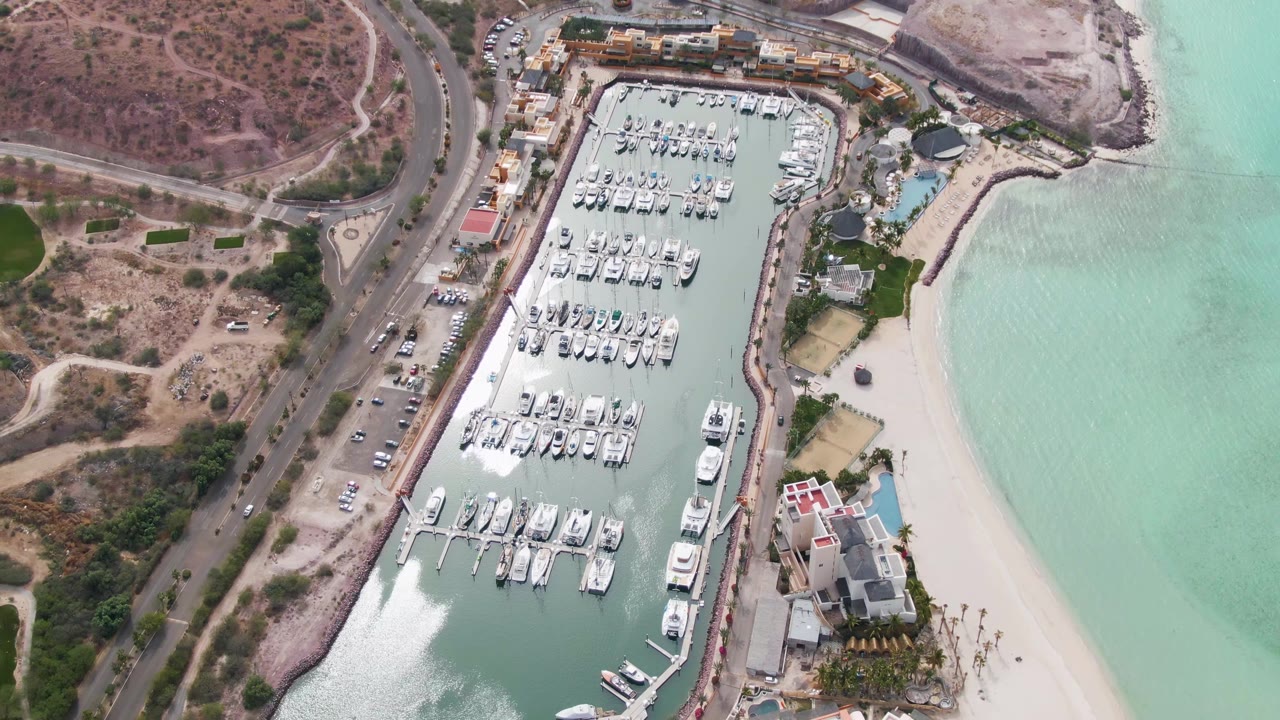 Aerial view over La Paz, Baja California Sur showing a marina packed with docked boats, a curved coastal road, pale beach sand, turquoise water, and no visible people at the opening moment.
