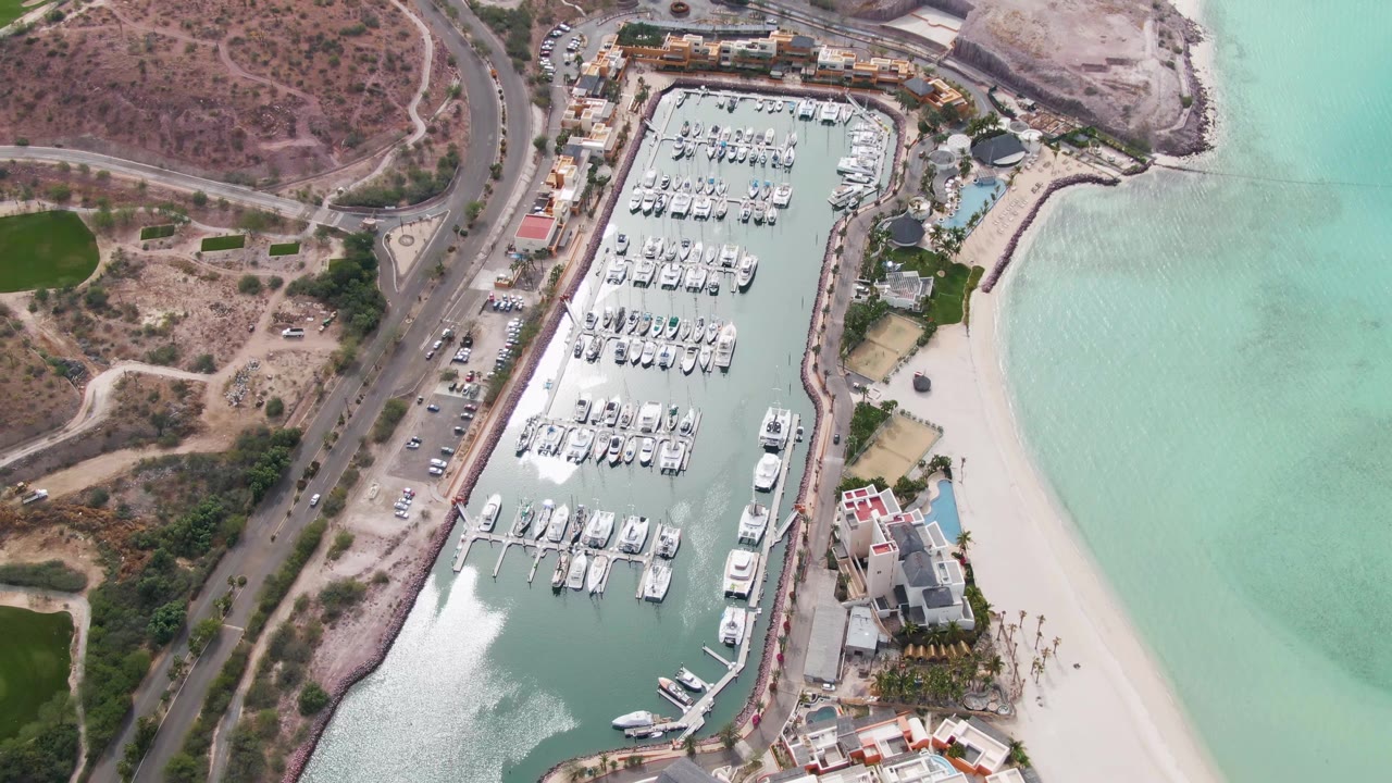 Top-down La Paz marina frame with rows of docked boats, cars on the adjacent road, a narrow sand beach, green-turquoise water, shoreline buildings, and no visible people.