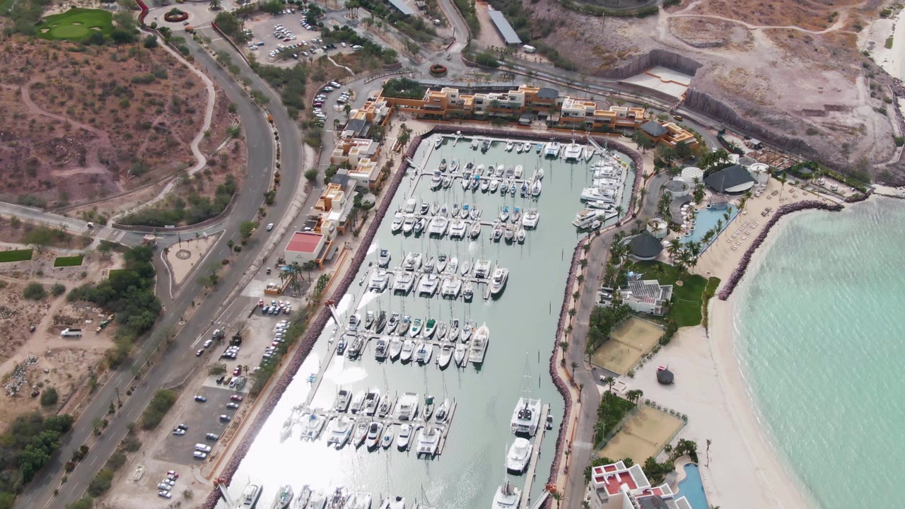 Aerial frame over La Paz, Baja California Sur showing the marina slightly farther along the shoreline, with dock rows centered, road traffic to the left, beach water to the right, and no visible people.