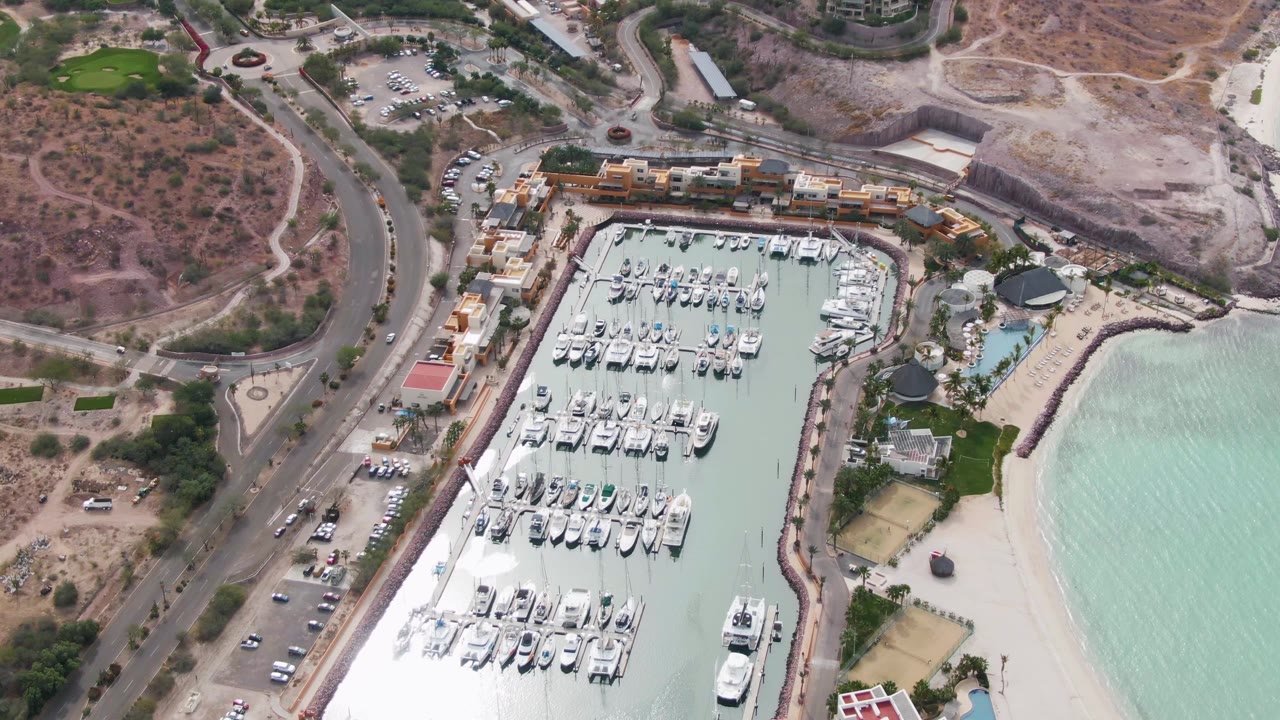 Mid-clip aerial view of La Paz, Baja California Sur with packed marina slips, a curved road and parked vehicles at left, beach and calm turquoise water at right, and no visible people.