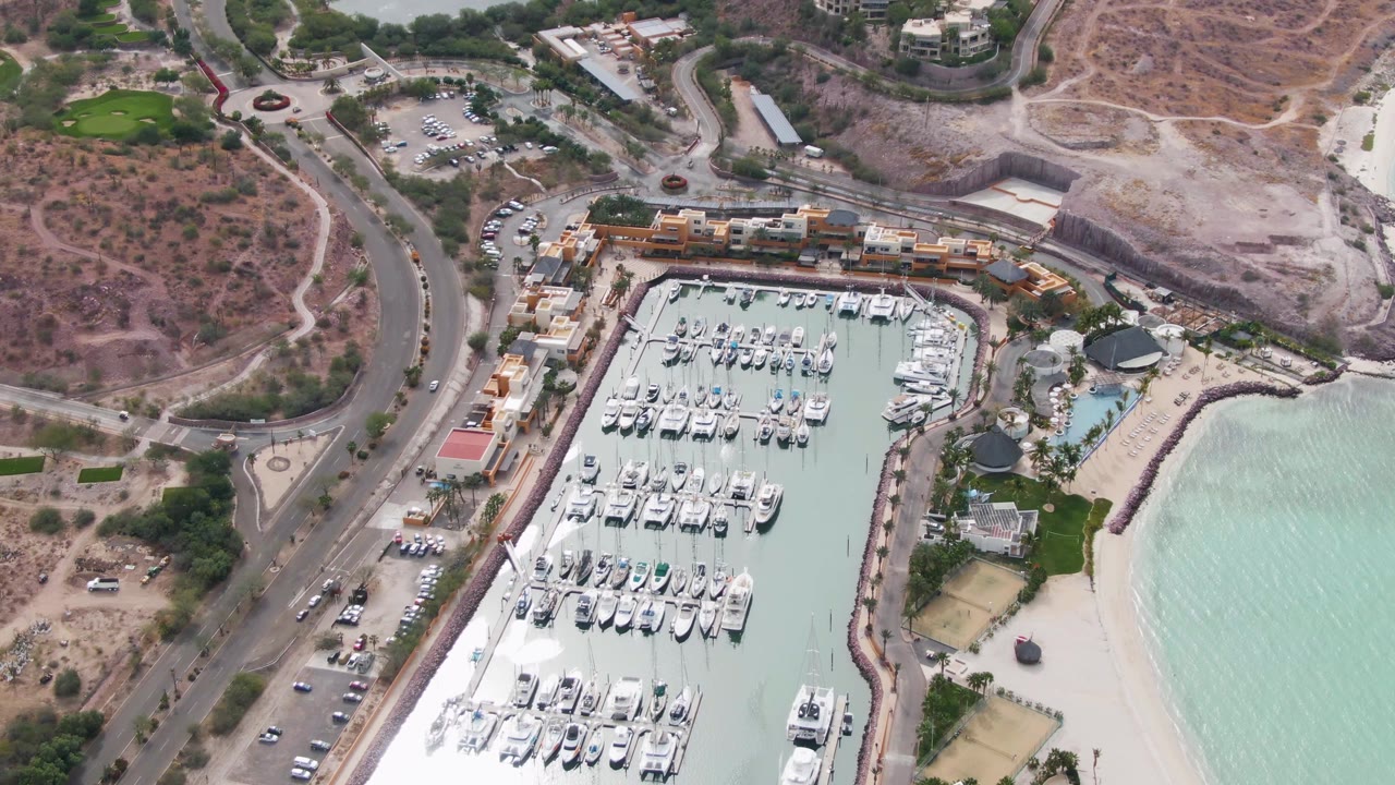 Aerial frame over La Paz, Baja California Sur with the marina shifted lower in view, the road and vehicles more prominent on the left, dry hillside above, beach water at right, and no visible people.