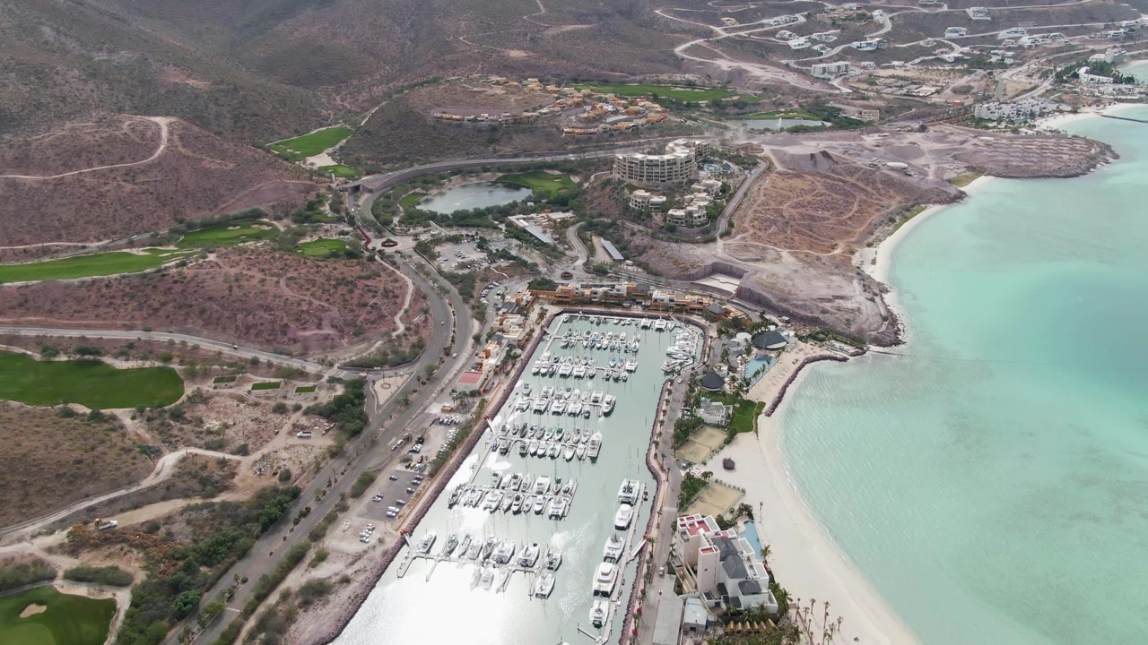 Aerial view over La Paz, Baja California Sur, with a docked-boat marina below, a pale beach beside turquoise water, dry hills, winding roads, green patches, and no visible people.