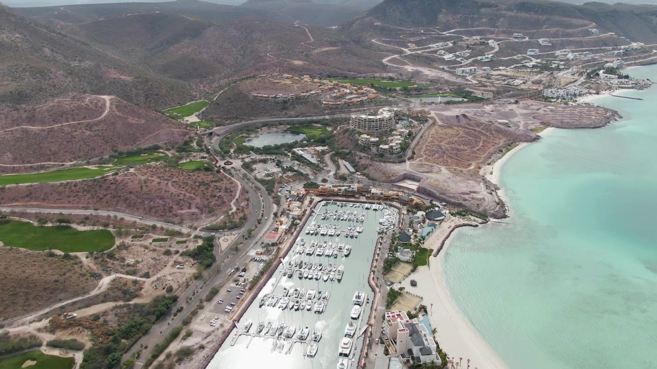 High aerial frame of La Paz marina with rows of white boats, a narrow sandy beach, turquoise bay water to the right, dry hillside roads beyond, and no visible people.