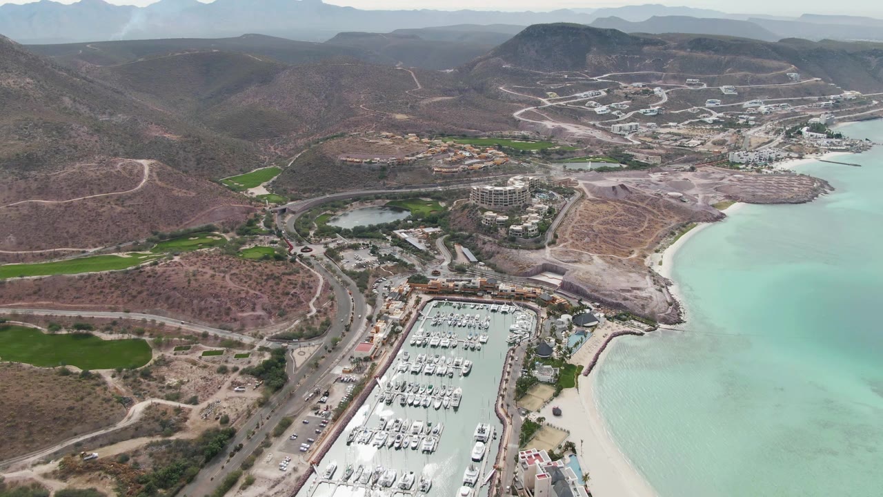 Wide La Paz aerial showing the marina lower in frame, turquoise water along the right edge, winding roads across dry hills, scattered buildings, distant mountains, and no visible people.