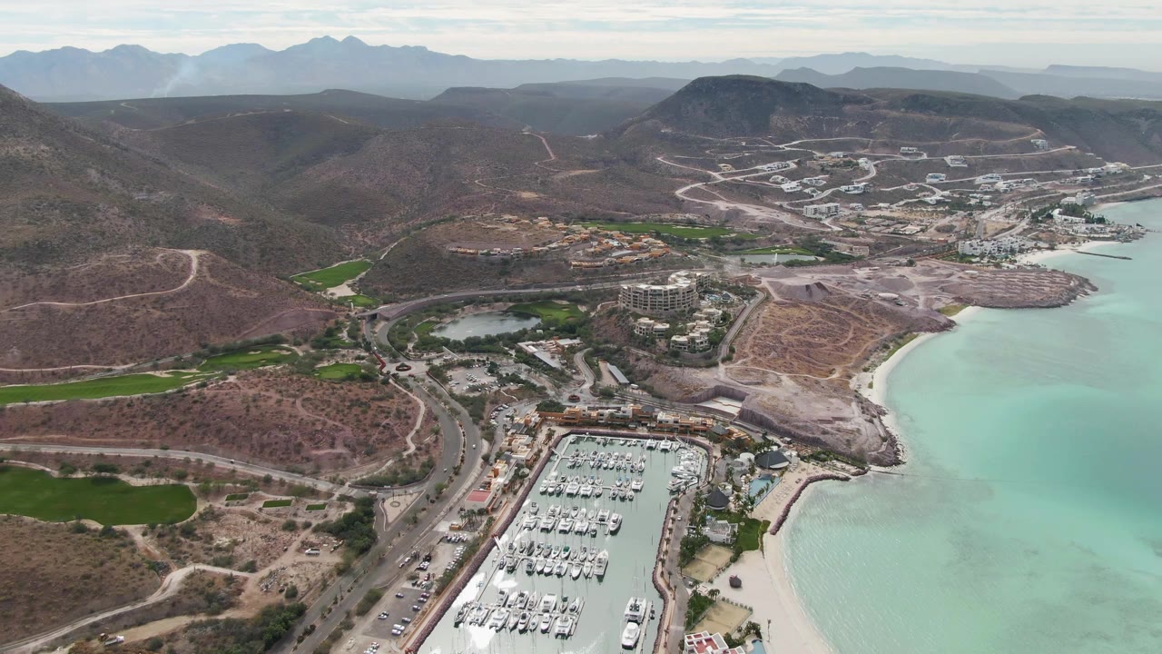 Aerial coastline view with marina, beach, roads, hills, and distant mountains.