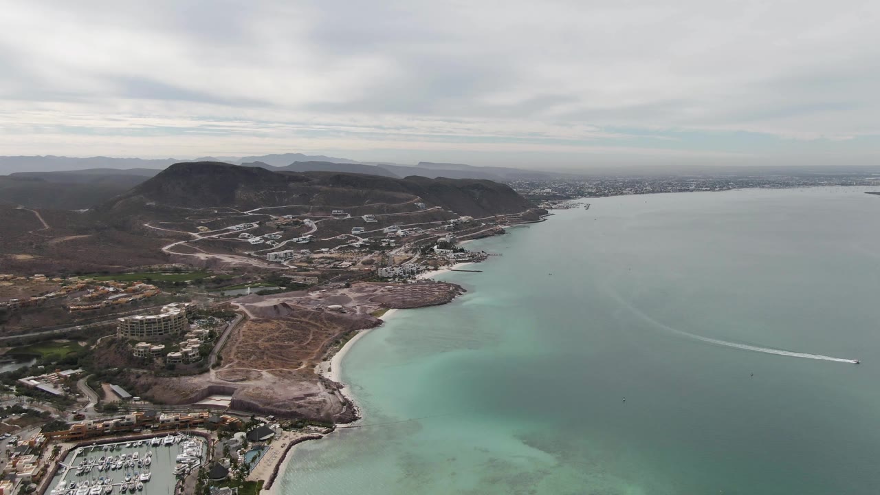 Aerial frame over La Paz, Baja California Sur showing the marina at lower left, a sandy beach curve, pale turquoise shallows, brown hills, distant city shoreline, a thin boat wake, and no visible people.