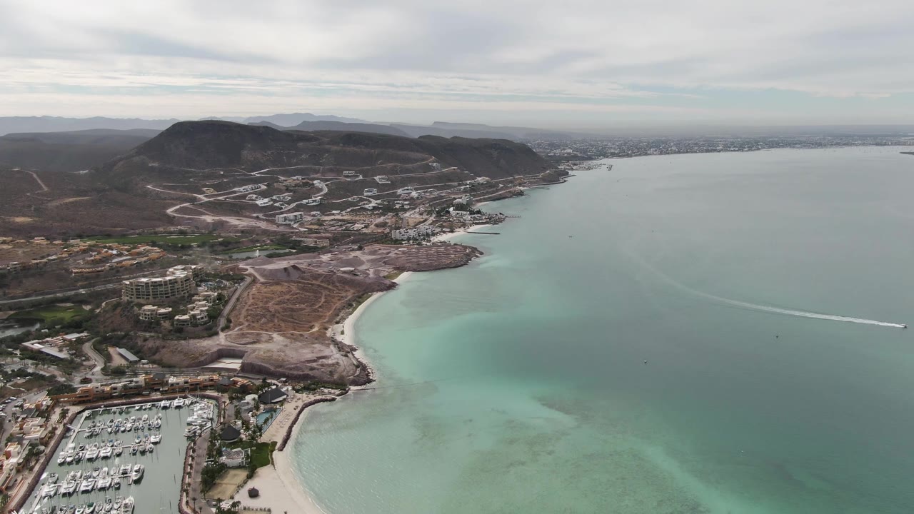 Aerial frame over La Paz showing more of the marina basin at the lower left, the pale beach arc, mottled turquoise shallows, darker bay water, hillside development, distant shoreline, and no visible people.