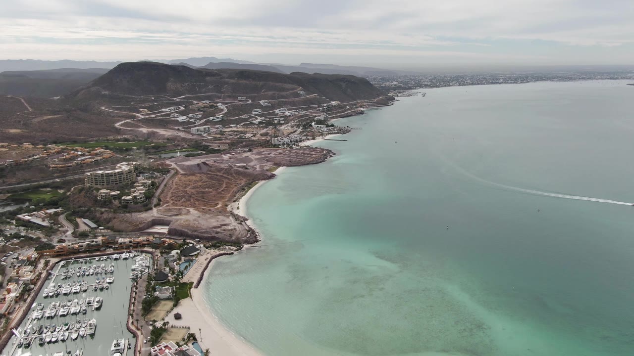 Aerial coastline view with marina, beach, turquoise shallows, hills, and wake line.