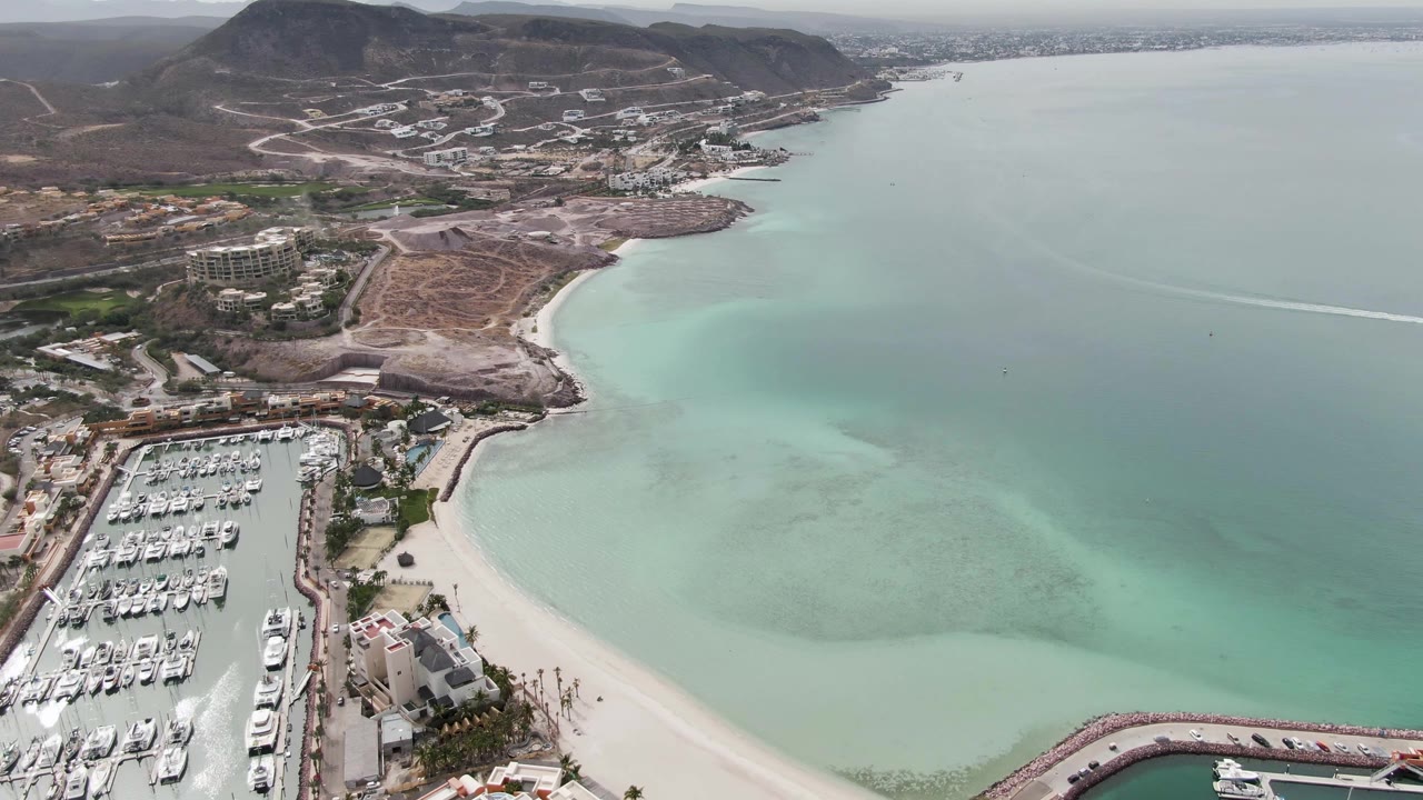 Opening aerial frame over La Paz, Baja California Sur, showing a marina full of white boats beside a curved sandy beach, pale turquoise water, dry hills, and no visible people.