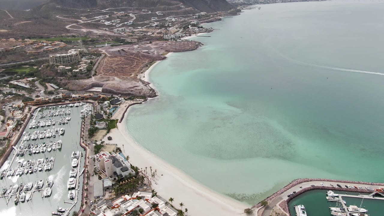 Aerial view in La Paz BCS with the marina shifted lower left, a broad curved sandy shoreline, shallow turquoise water with darker patches, hillside development, and no visible people.