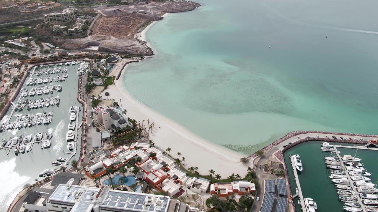 High aerial view of marina slips, beach curve, clear shallow water, and hillside shoreline.