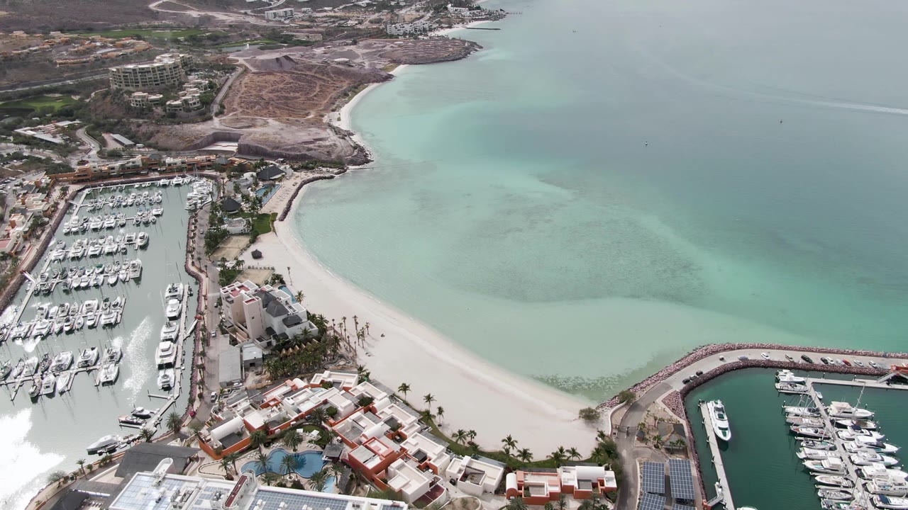 Drone view over La Paz, Baja California Sur showing rows of boats in the marina, a pale sand beach, broad turquoise bay water, distant shoreline, brown hills, and no visible people.