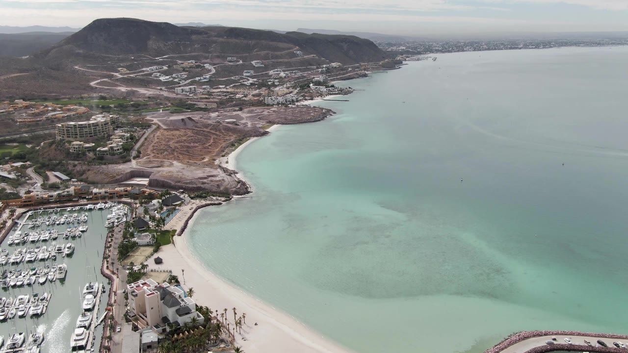 High aerial view in La Paz, Baja California Sur, showing a marina full of white boats at left, a curved pale beach, turquoise bay water, dry hills, roads, and no visible people.