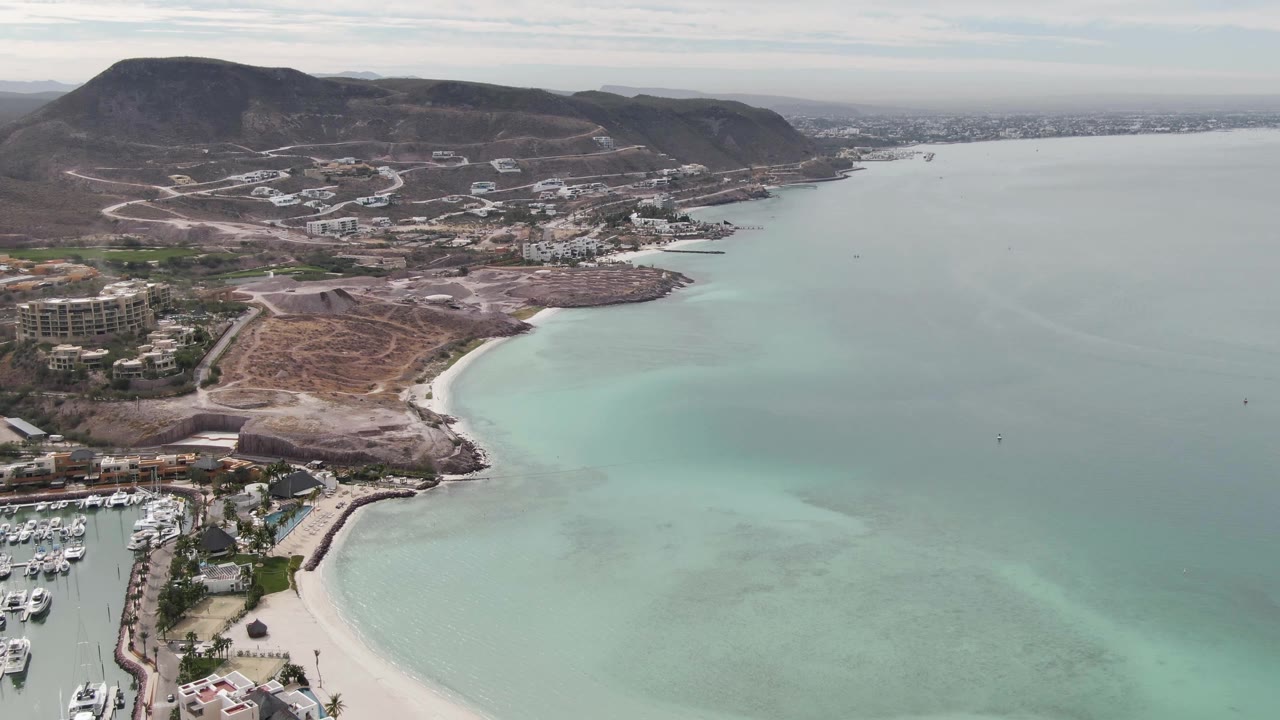 Aerial frame from La Paz showing the marina partly cropped at left, a small sandy cove and rocky point below, translucent turquoise shallows, dry coastal hills, roads, and no visible people.