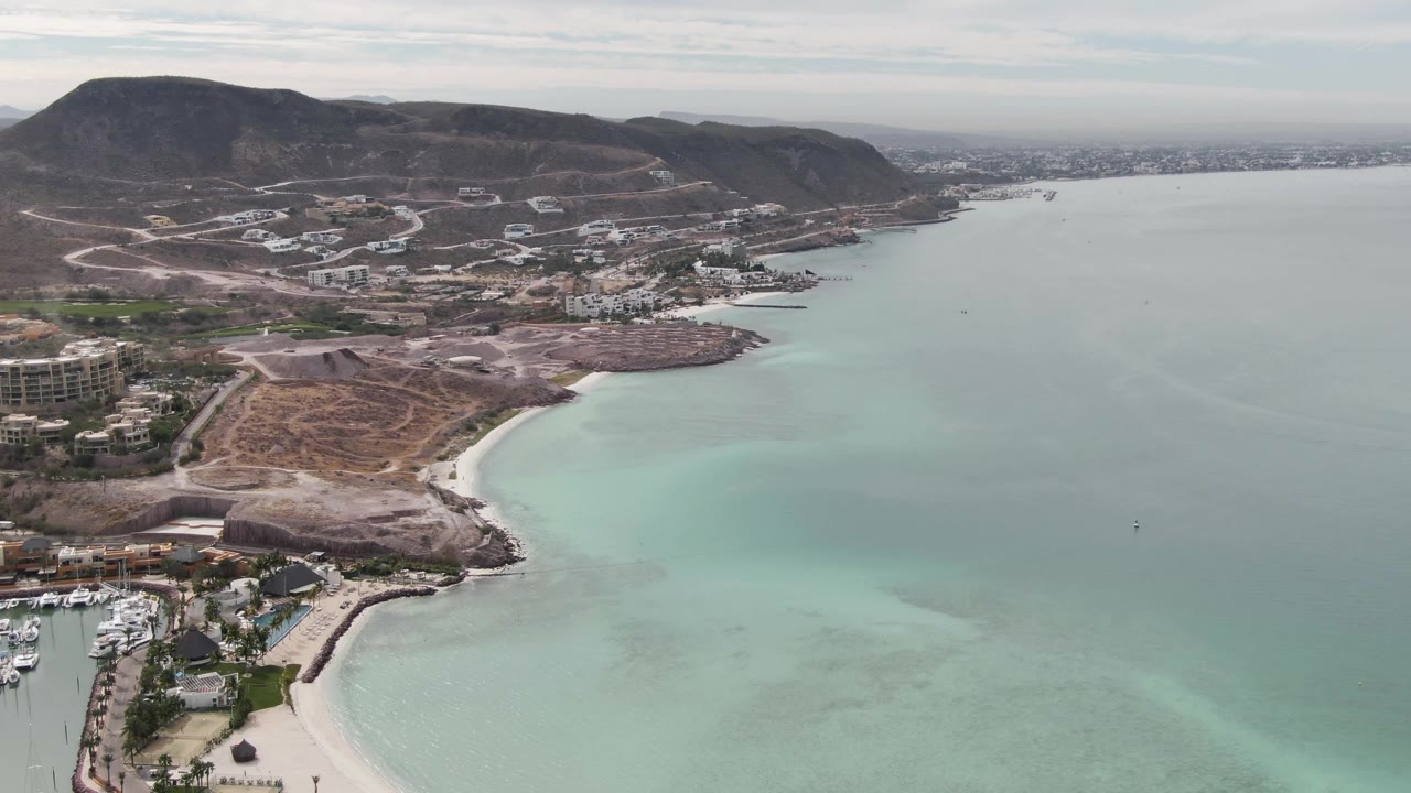 La Paz aerial shoreline frame with most of the marina nearly out of view at lower left, a pale beach curving along the bay, soft turquoise water gradients, hillside construction roads, and no visible people.