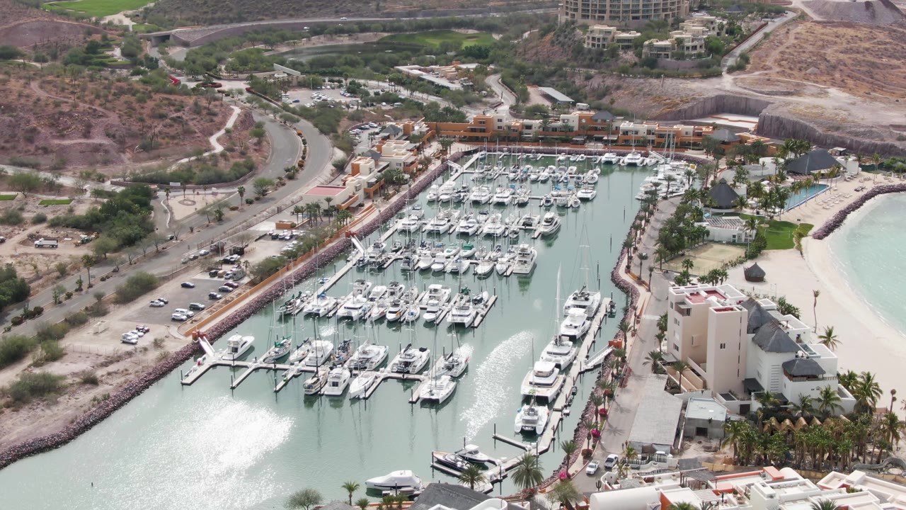 Overhead La Paz marina frame with white boats filling the center slips, a coastal road and scattered cars to the left, pale beach water to the right, hillside development in the distance, and no visible people.