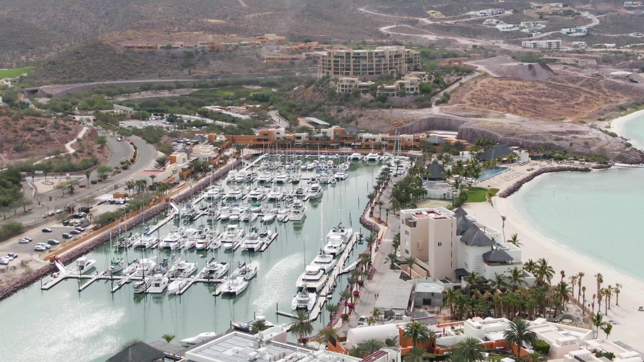 A second aerial frame shows the La Paz marina centered more tightly, with boat slips, a pale sand beach to the right, palm-lined waterfront structures, roads, hills, and no visible people.