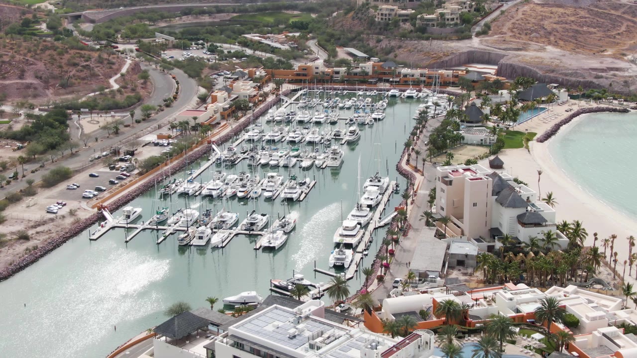 Aerial view of the La Paz marina as a bright vessel wake appears near the lower-left water, with docked boats, beach, palms, buildings, and no visible people.