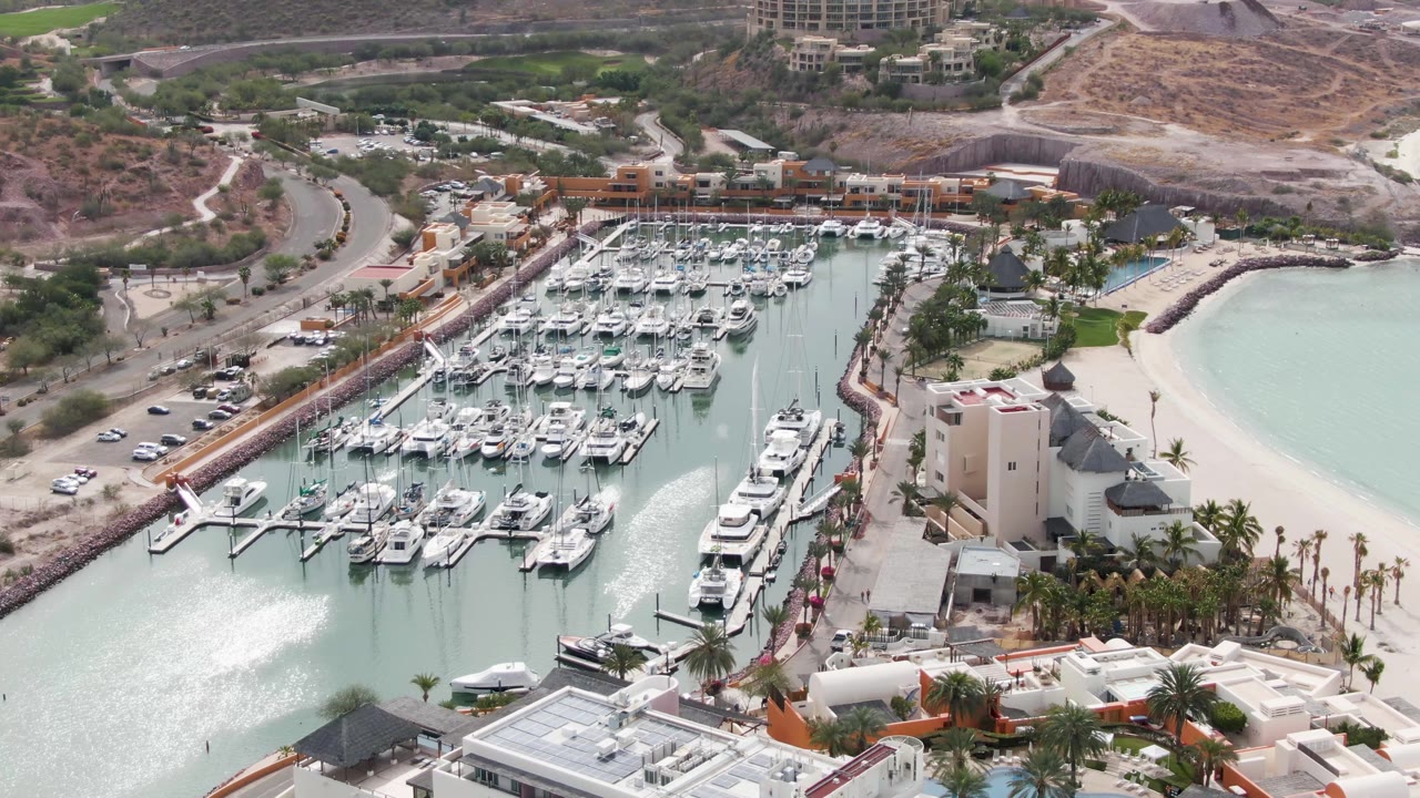 La Paz marina aerial with the white wake spreading farther across the left side of the water, while docked boats, a sandy beach, palm trees, and hillside roads remain visible with no visible people.