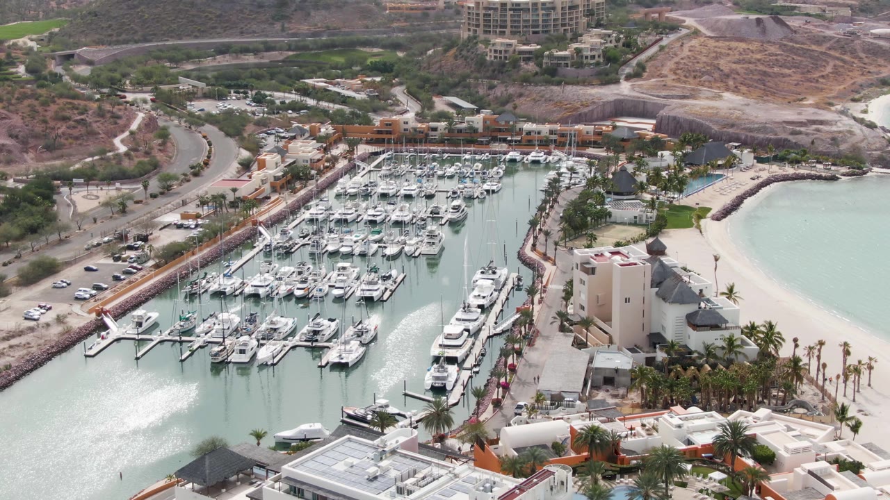 High aerial frame over the La Paz marina with the wake now brighter across the lower-left water, rows of docked boats in the center, beach and buildings to the right, and no visible people.
