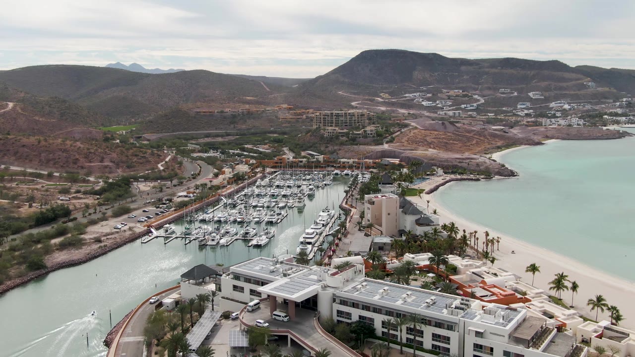 Aerial frame from La Paz, Baja California Sur showing a pale canal beside a crowded marina, white waterfront buildings, a sandy beach, arid hills, and no visible people.
