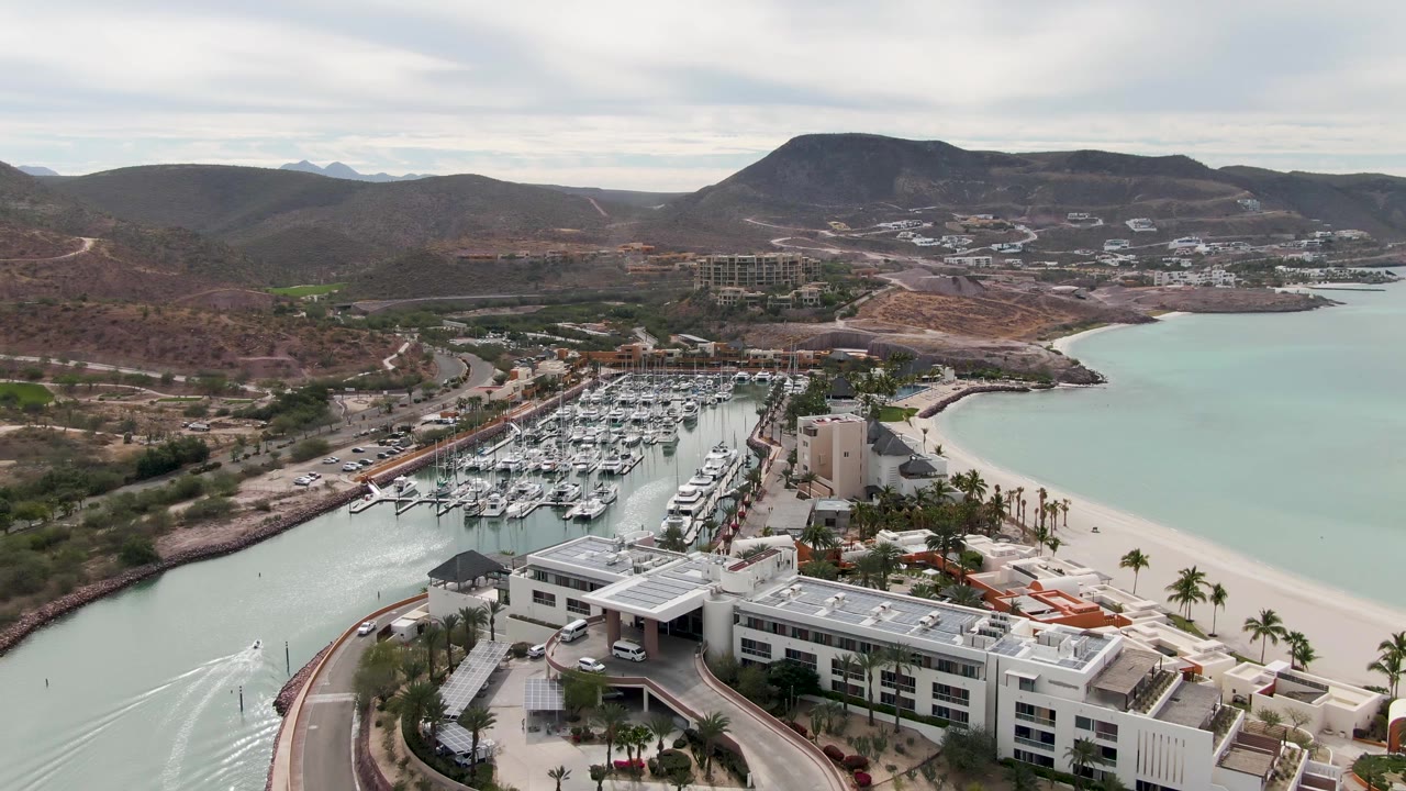 Aerial view in La Paz, Baja California Sur with foreground white buildings, a curved waterfront road, marina slips, pale canal water, beach, hills, and no visible people.