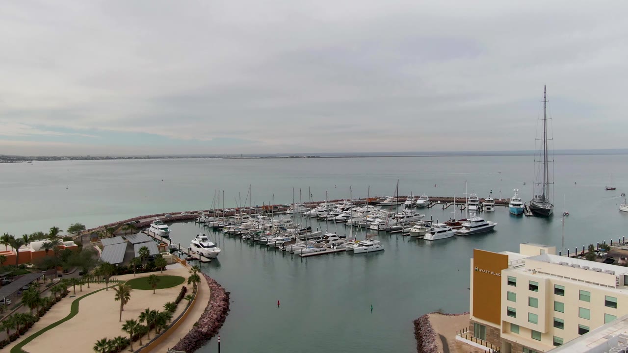Aerial frame in La Paz showing the marina basin slightly farther across the docks, with clustered boats, open bay water, palm trees along the walkway, a waterfront building, and no visible people.