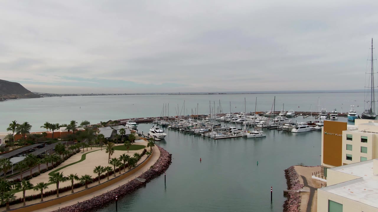 Opening frame of a La Paz, Baja California Sur marina with docked boats across calm gray-green water, a palm-lined waterfront road, a building edge at right, distant hills, and no visible people.
