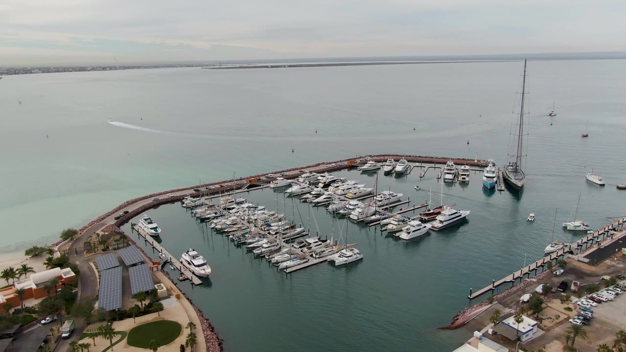Aerial view in La Paz showing marina slips filled with white boats, pale green-blue water, a long breakwater, distant shoreline, and no visible people.