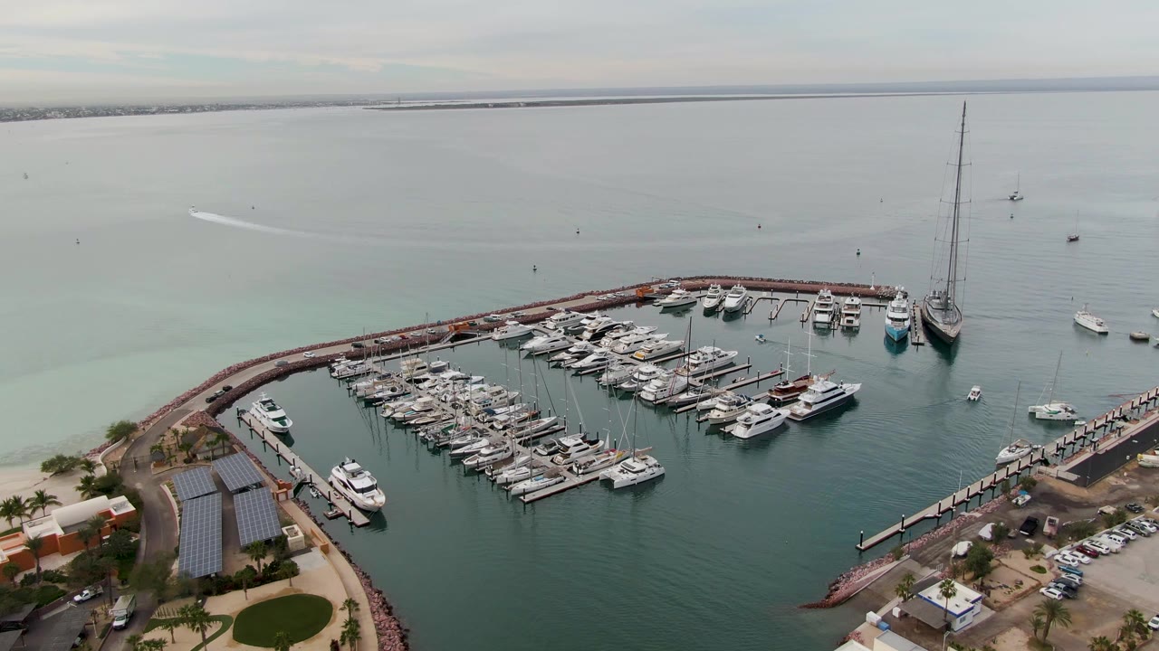 Wide aerial frame over the La Paz marina with docked yachts, a tall sailboat mast near the right edge, open water, and no visible people.