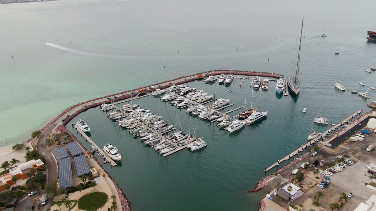 At 0:02, the La Paz marina aerial shows dense boat slips inside the curved breakwater, small boats scattered offshore, hazy gray-green water, and no visible people.