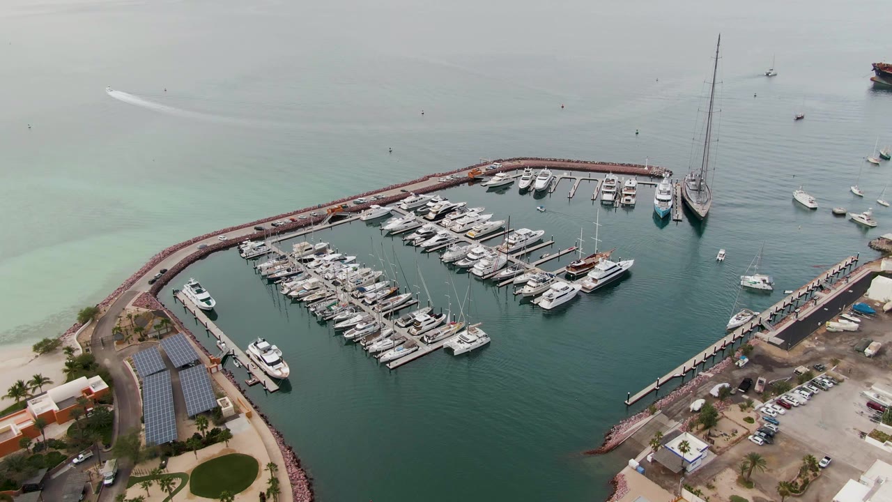 At 0:03, the La Paz marina is seen from above with the curved breakwater, tightly packed docks, a right-side pier, shoreline parking, solar panels, and no visible people.